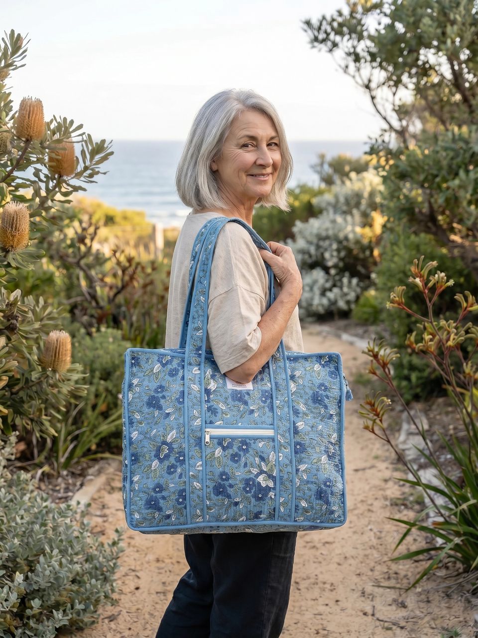 Woman holding a blue floral tote bag in a natural setting with plants and a path.